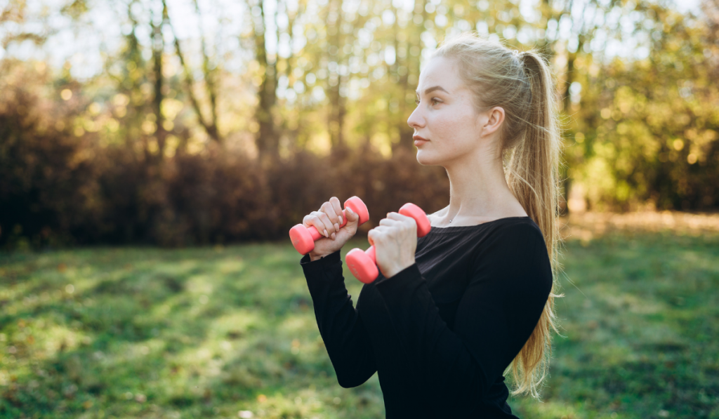 Profile of slim girl with dumbbells outdoors. Fitness in park.